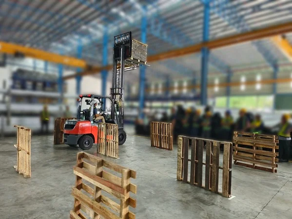 An operator maneuvers a red forklift lifting a pallet during a hands-on forklift training session in a warehouse.